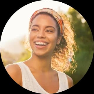A joyful young woman with curly hair wearing a headband smiles in the sunlight.